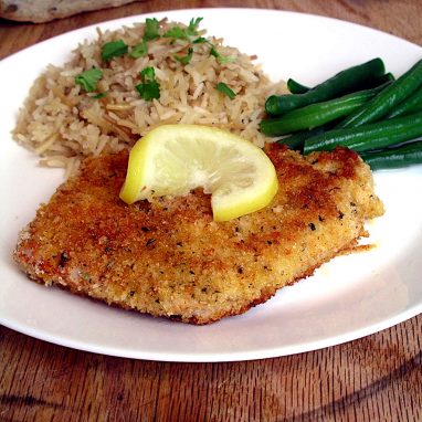 a breaded pork cutlet on a white plate with rice pilaf and green beans